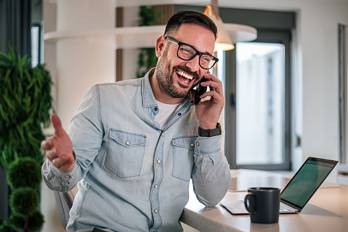 Experienced businessman smiling during a phone call Happy young adult businessman communicating with his business partners over the phone Successful entrepreneur working in a modern workspace