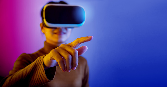 Asian woman wearing VR glasses pointing finger to do activities in the virtual world on blue and purple background.