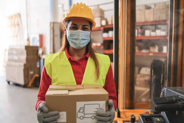 istock Hispanic warehouse worker woman holding delivery box while wearing safety mask - Focus on face 1368007620