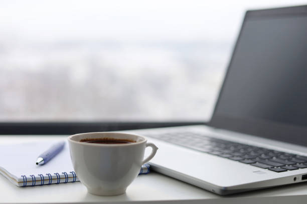 istock Coffee cup, laptop and notepad with pen on a table against the window, view to city 1368011669
