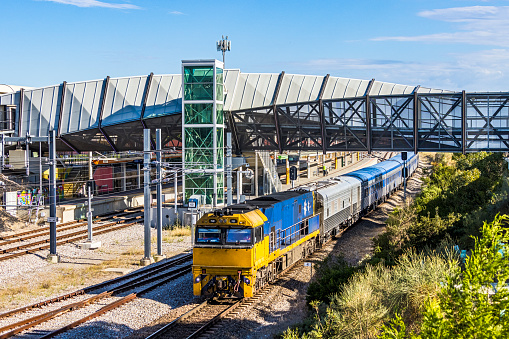 Australian Overland Passenger Train passing modern metro train station. The train provides interstate links to Eastern and Western State rail services as well as North-South luxury trains. ID, logos, murals edited.