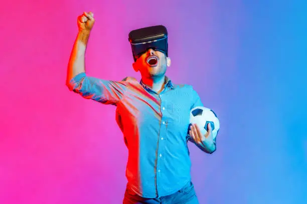 Photo of Man in headset and holding soccer ball, watching 3d football team, cheering, celebrating victory.