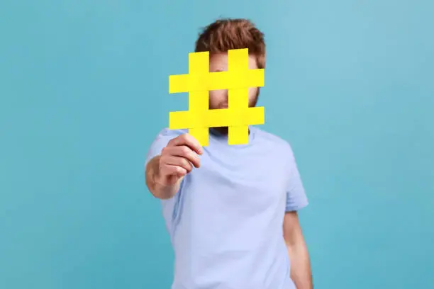 Photo of Man in T-shirt hiding behind yellow hashtag symbol covering face with hash sign, concept of blogging