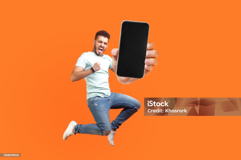 Happy man flying and jumping in air and showing big mobile empty screen Happy man flying and jumping in air and showing big mobile empty screen for copy space and advertising area. indoor studio shot isolated on orange background Mobile Phone Stock Photo