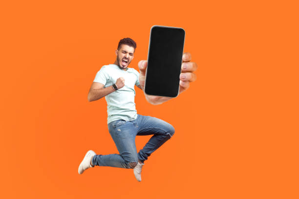 Happy man flying and jumping in air and showing big mobile empty screen Happy man flying and jumping in air and showing big mobile empty screen for copy space and advertising area. indoor studio shot isolated on orange background air jump stock pictures, royalty-free photos & images