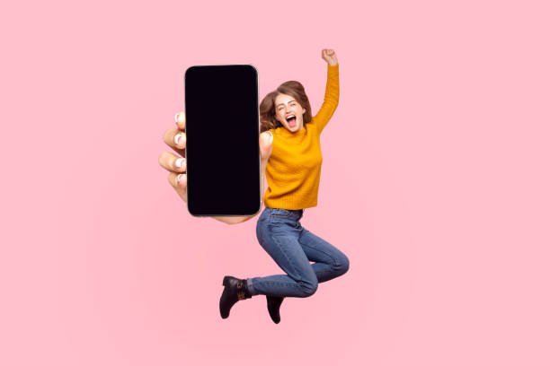 Happy young woman flying and jumping in air and showing big mobile empty screen Happy young woman flying and jumping in air and showing big mobile empty screen for copy space and advertising area. indoor studio shot isolated on pink background human representation photos stock pictures, royalty-free photos & images