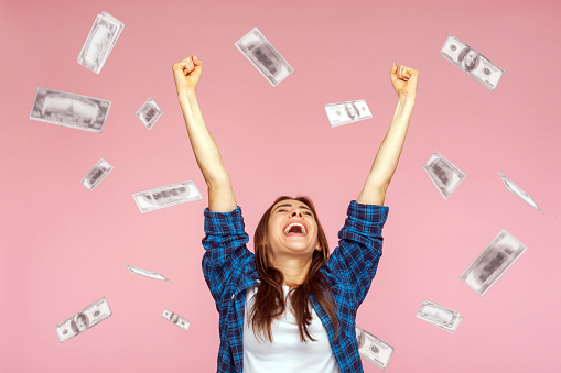 Money rain, winner and rich. Hurray. satisfied happy young woman rising hands up with toothy smile on face, pleased, amazed with money falling. Indoor studio shot isolated on pink background