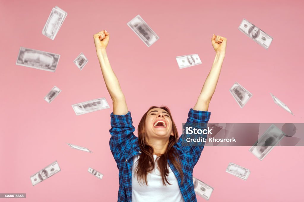 Money rain, woman rising hands up with toothy smile on face, pleased, amazed with money falling. Money rain, winner and rich. Hurray. satisfied happy young woman rising hands up with toothy smile on face, pleased, amazed with money falling. Indoor studio shot isolated on pink background Currency Stock Photo