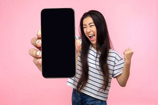 Portrait of euphoric happy joyful girl with long hair screaming with happiness showing mobile advertisement mockup area and celebrating her victory. indoor studio shot isolated on pink background