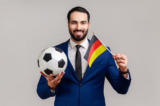 Positive bearded man holding flag of germany and soccer black and white classic ball and watching match, wearing official style suit. Indoor studio shot isolated on gray background.