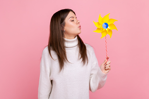 Portrait of carefree brunette woman blowing at paper windmill, playing with pinwheel toy on stick, wearing white casual style sweater. Indoor studio shot isolated on pink background.