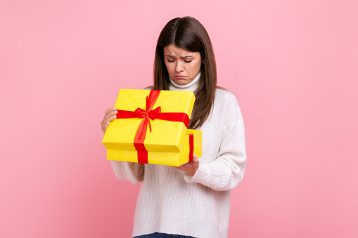 Unhappy dark haired girl looking inside wrapped present box, open gift, expressing disappointment, wearing white casual style sweater. Indoor studio shot isolated on pink background.