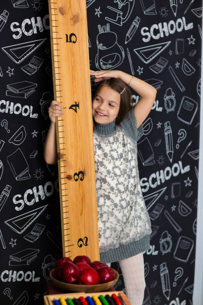 une fille en robe grise se tient près d’une grande règle. elle mesure sa taille. elle va à l’école - schoolgirl child backpack book bag photos et images de collection