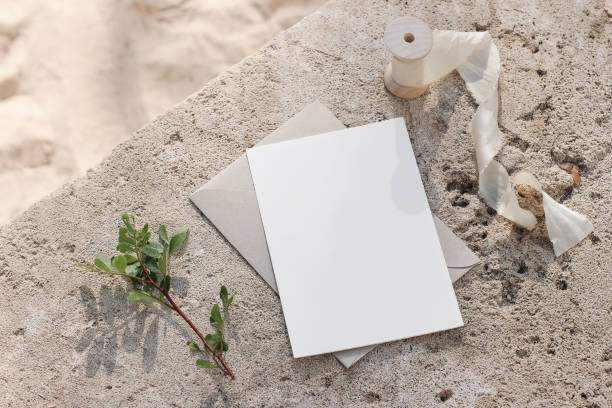 Summer wedding stationery mock-up. Blank greeting card, invitation. Envelope and silk ribbon in sunlight. Green lentisk branch. Beige concrete background in sunlight. Blurred sandy beach. Top view. stock photo