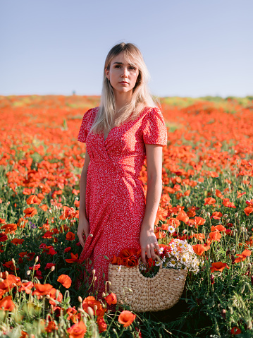 Caucasian blonde woman in a poppy field. Concept of freedom with soft colors. Woman in red dress and handbag with wild flowers