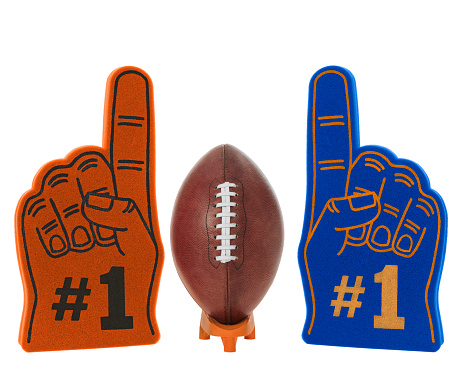 A brown leather American football with white laces sitting on a kicking tee along with a large Orange and Black foam #1 finger on one side and a blue and yellow foam #1 finger on the other side, team colors for each of the teams playing in the BIG GAME, on a white background.