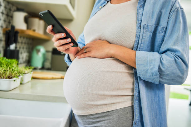 Cropped shot of an unrecognisable pregnant woman standing alone in her kitchen and using her cellphone Is what I'm feeling normal? Bloating stock pictures, royalty-free photos & images