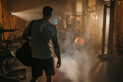 Fit man carrying gym bag walking in dark gym filled with smoke alone.