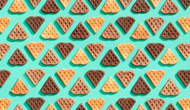 Many heart-shaped waffles aligned on a green-colored background, view from above. Pattern with delicious waffles made with cacao, vanilla, and cinnamon.