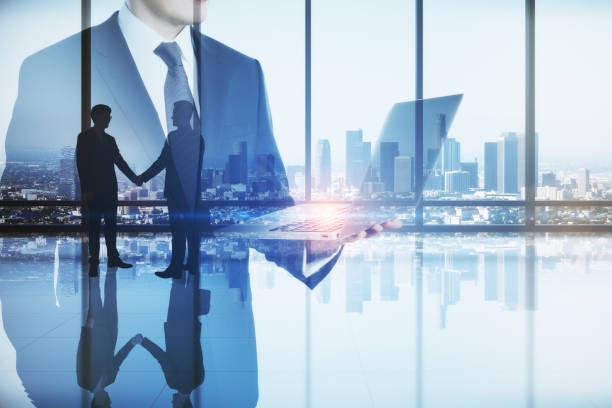 istock Abstract image of businessman using laptop computer in modern office interior with panoramic city view, businesspeople shaking hands and mock up place. Teamwork, leadership and technology concept. Double exposure. 1368053939