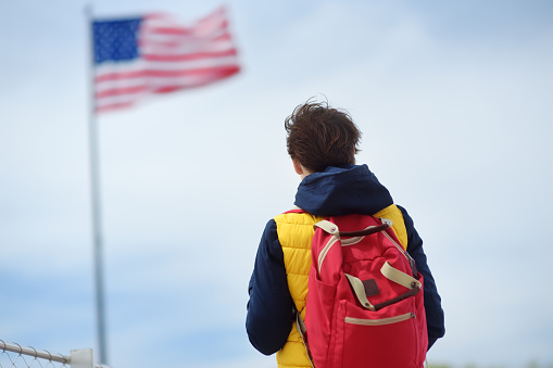 Tourist is traveling to Island of liberty. New York, USA. Woman with backpack is on the background of blue sky and fluttering of American flag. Sightseeing of usa.