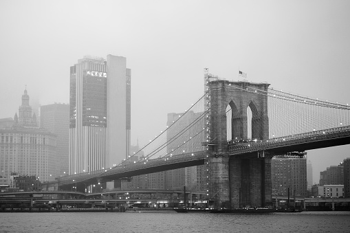Black and white photo of skyscrapers of Manhattan and Brooklyn bridge on foggy and cloudy day. Famous bridge. Postcard view of New York. Travel, tourism, sightseeing of USA.