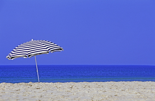 Side view of elegant blonde woman with sun hat looking at blue-white parasols on French Riviera sea beach. Copy space