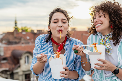 Photo of two female friends on the rooftop of the building eating fast food, and laughing