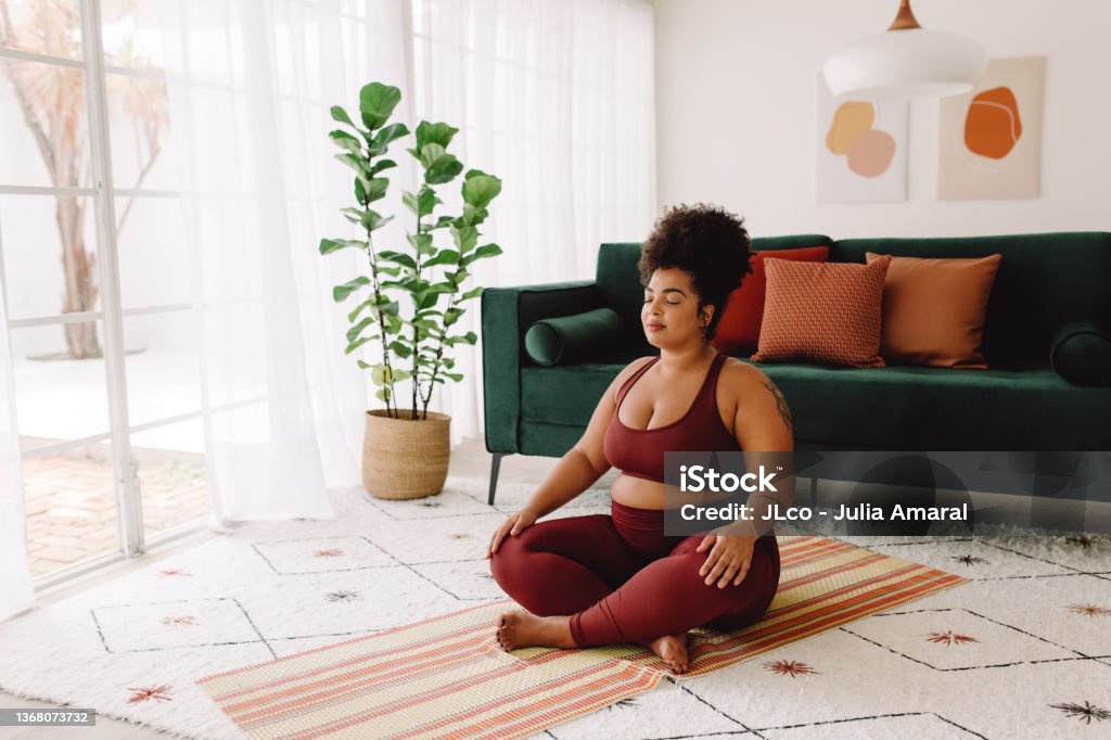 Plus size woman practicing meditation at home Body positive woman doing mindfulness workout at home. Beautiful female in sports wear practicing meditation at home. Meditating Stock Photo