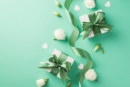 Top view photo of two small white gift boxes tied up with green silk bow and scattered eustomas flower buds and confetti in shape of hearts on the pastel turquoise background