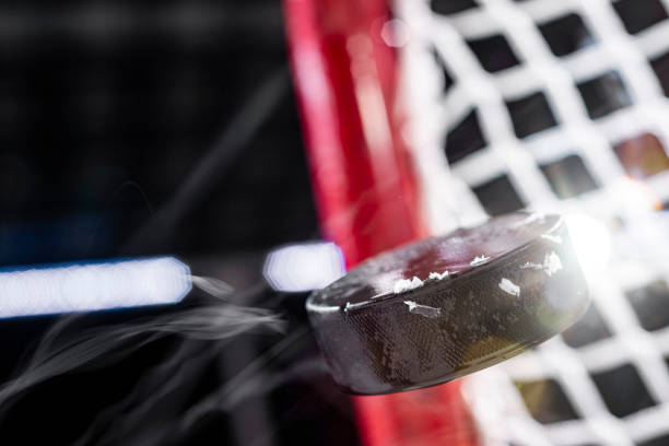 A close-up of an ice hockey puck in mid-air of a goal net with flare from the arena lights Looking up at an ice hockey puck flying through the air with ice shavings as it heads to the goals net, with flare from the arena lights. ice hockey net stock pictures, royalty-free photos & images