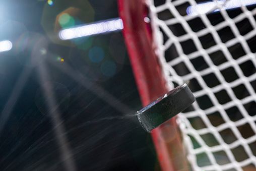 Looking up at an ice hockey puck flying through the air with ice shavings as it heads to the goals net, with flare from the arena lights.