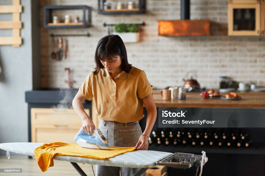 Asian Young Woman Ironing Clothes Waist up portrait of young Asian woman ironing clothes at home and doing household chores, copy space Iron - Appliance Stock Photo