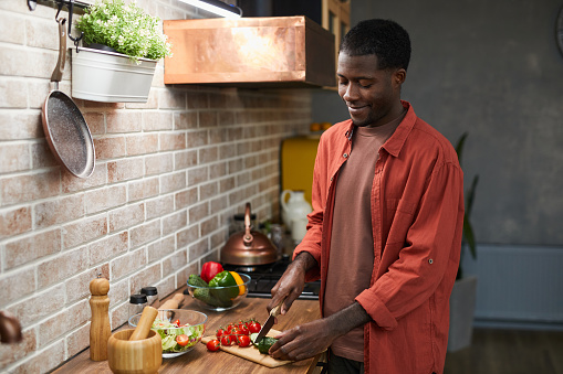 Portrait of smiling black man cutting vegetables while cooking in cozy kitchen, copy space