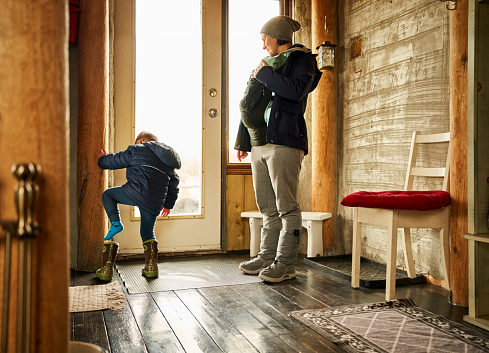 Small boy putting on his boot with his mother standing by carrying a baby boy at front door of eco cabin