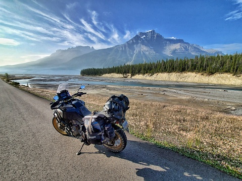 A motorcycle sits on a gravel road overlooking mountains and a river valley along the Icefields Parkway. A light wildfire haze is visible. Banff National Park, Alberta, Canada