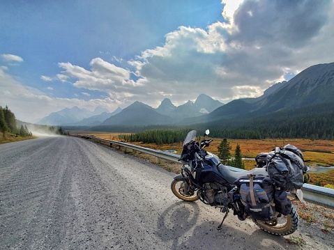 A motorcycle sits on a gravel road overlooking mountains and a golden valley. A light wildfire haze is visible. Peter Lougheed Provincial Park, Kannanaskis Country, Alberta, Canada