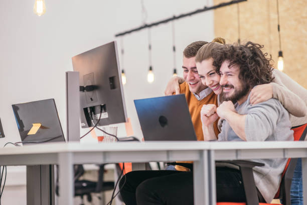 happy surprise for the teammates. coworkers cheering while looking at a computer, screen at the office. positive vibe at the startup modern office. - cheering business three people teamwork imagens e fotografias de stock