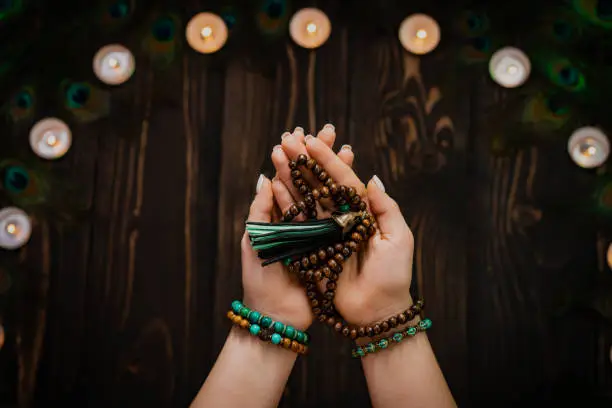 Woman holds in hand wooden mala beads strands used for keeping count during mantra meditations. Weaving and creation. Wooden background with candles and feathers. Spirituality, religion, God concept