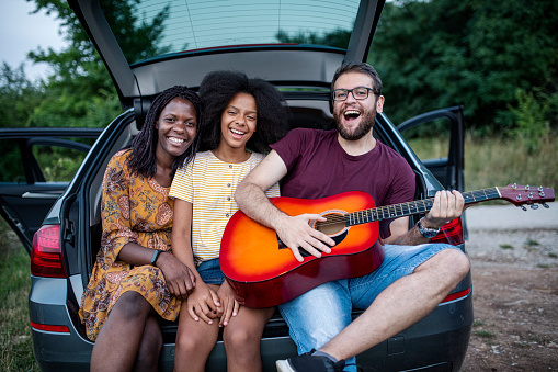 Portrait of happy diverse family sitting on car trunk and playing guitar