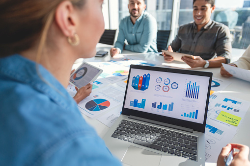Multi racial group of people working with Paperwork on a board room table at a business presentation or seminar. The documents have financial or marketing figures, graphs and charts on them. There is a laptop with graph and chart infographic data
