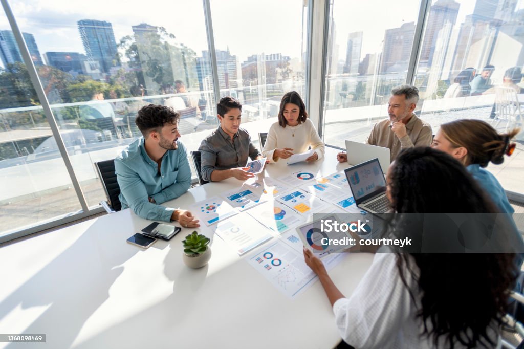 Multi racial group of people working with Paperwork on a board room table at a business presentation or seminar. Multi racial group of people working with Paperwork on a board room table at a business presentation or seminar. The documents have financial or marketing figures, graphs and charts on them. There is a laptop and digital tablet  on the table. Multi ethnic group including Caucasian and African American Data Stock Photo
