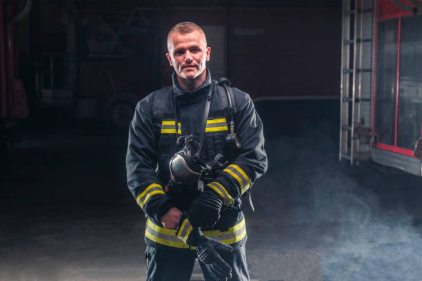 retrato de um bombeiro usando bombeiros e capacete. fundo escuro com fumaça e luz azul. - turnouts - fotografias e filmes do acervo