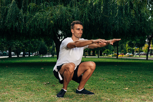 Image of a handsome cheerful young sports fitness man standing in green park nature make sport exercises for legs squats