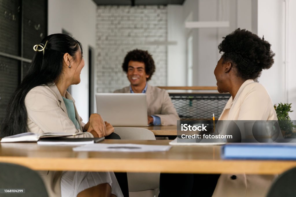 Two young businesswomen casually chatting with their male colleague Front view of two cheerful young businesswomen sitting at the table, turning around and casually chatting with their male colleague sitting at his desk behind them. 25-29 Years Stock Photo