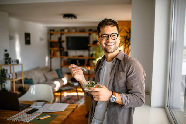 Healthy lunch at home Young millennial man at home, standing next to the window, smiling, having a breakfast. lunch break stock pictures, royalty-free photos & images