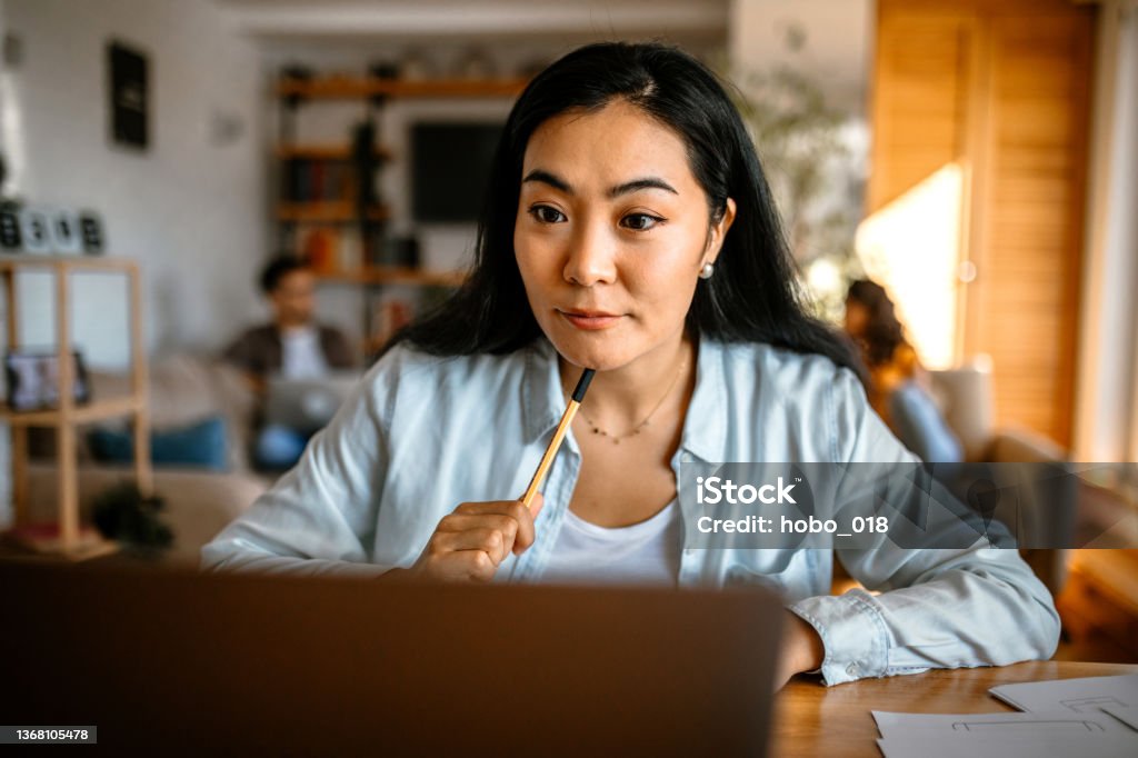 Young pretty asian woman sitting at the desk in her home office, doing home finances. Group of students studying at home. Young smiling asian woman working using laptop, while her friends working in background. E-Learning Stock Photo