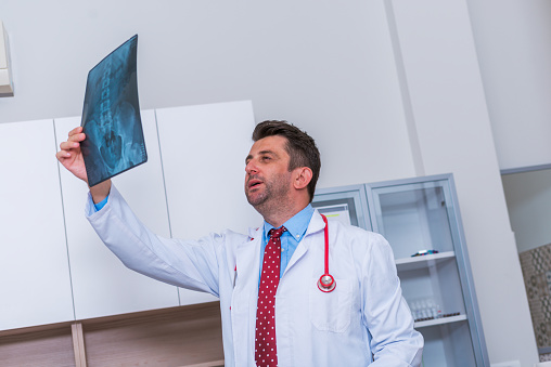Male Doctor in his mid 40's reading an x-ray image ( radiography ) from a patient hip region ( coxa ) while standing in a hospital radiology department.