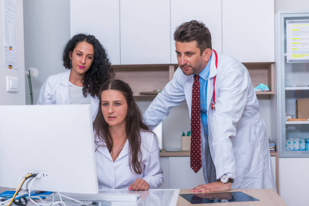 istock Two nurses ( female physicians ) and a male doctor standing next to a pc, mac ( desktop computer ) in the doctor's office while discussing a medical report. 1368105525