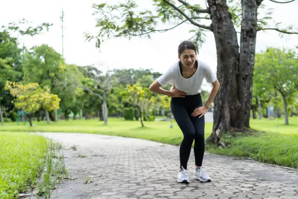 Photo of Asian attractive sport woman feels pain on stomach after run on street.Beautiful girl athlete in sportswear having stomach ache illness problem while exercise by jogging workout in outdoor public park